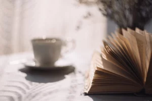photographie d'une partie d'un livre ouvert en premier plan, les pages défilent en statique et en second plan flou il y a une tasse de thé illuminé par une fenêtre. Cela crée une ambiance cosy.